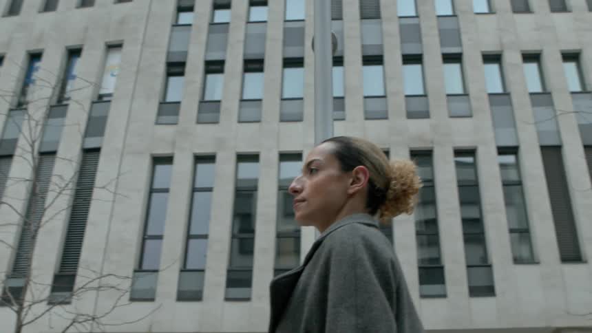 A woman in a gray coat stands in front of a modern building with tall narrow windows