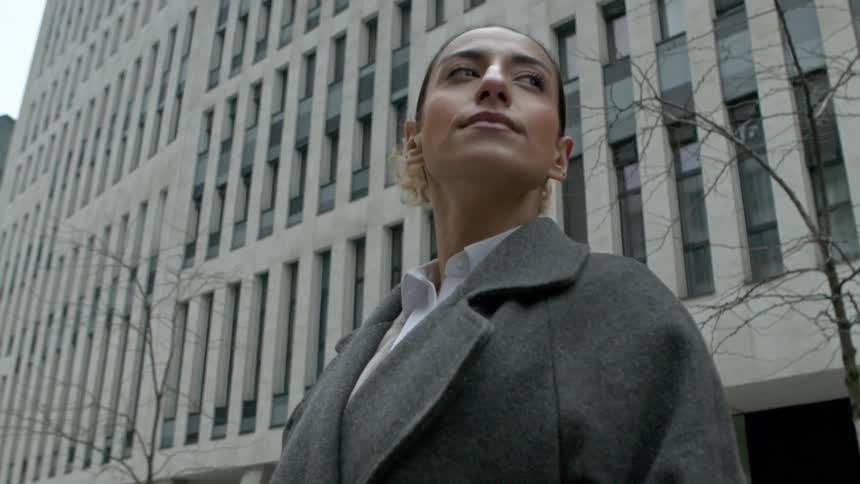 A woman in a gray coat stands in front of a large building and looks up