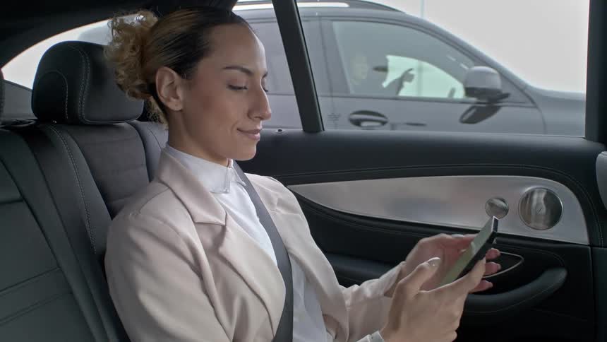 A woman is sitting in the backseat of a car wearing a lightcolored blazer and using a smartphone