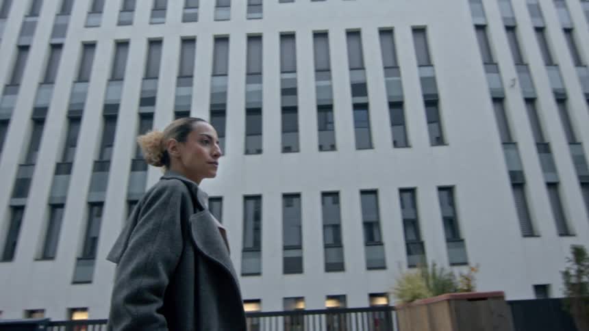 A woman wearing a grey coat and white shirt is walking between modern multistory buildings on a cloudy day