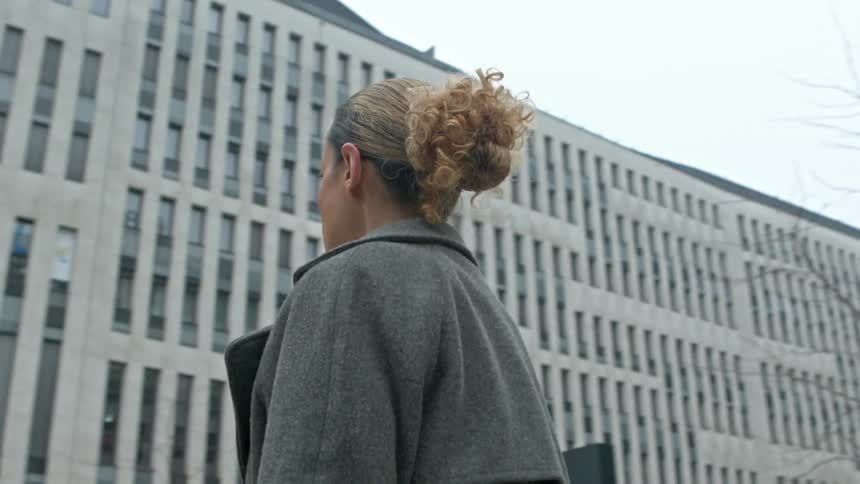A woman wearing a grey coat and white shirt is walking between modern multistory buildings on a cloudy day