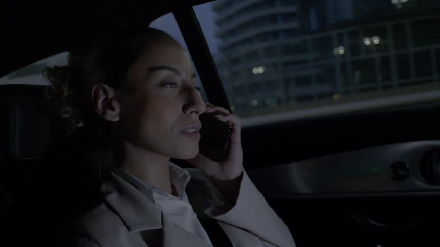 A young woman is sitting in the back seat of a car talking on her cell phone and staring straight ahead
