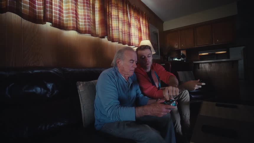 Father and son are sitting on a dark leather couch in a dimly lit living room playing video games