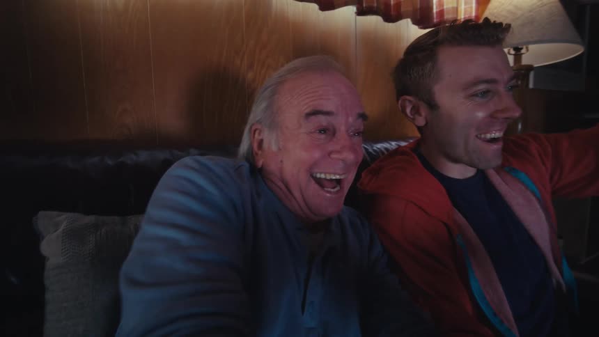 Father and son are sitting on a dark leather couch in a dimly lit living room playing video games