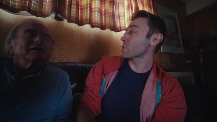 Father and son are sitting on a dark leather couch in a dimly lit living room playing video games