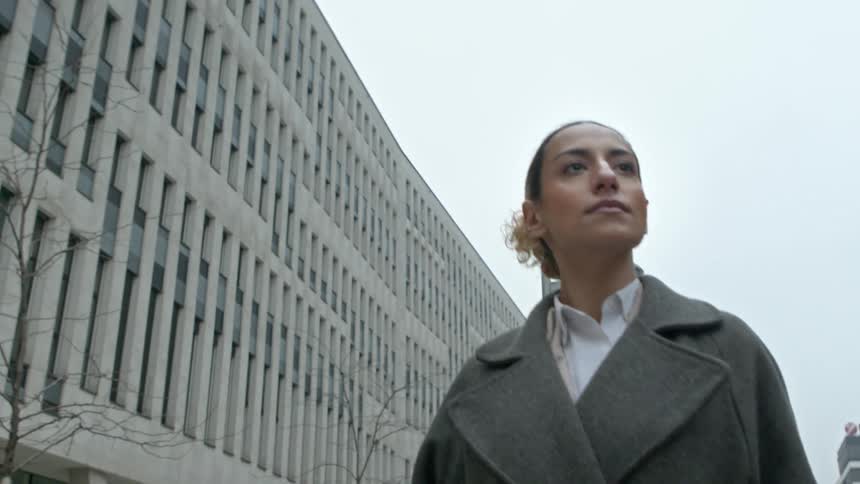 A person in a gray coat walks on a city street with modern multistory buildings with a cloudy sky overhead