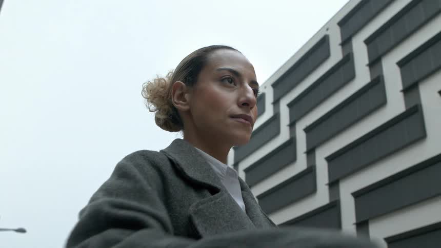 A young woman in a gray coat looking at a watch on her wrist and behind her there are some buildings