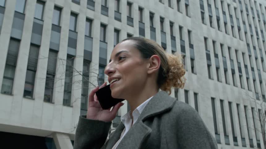 A woman in a coat talks on the phone while walking past modern city buildings