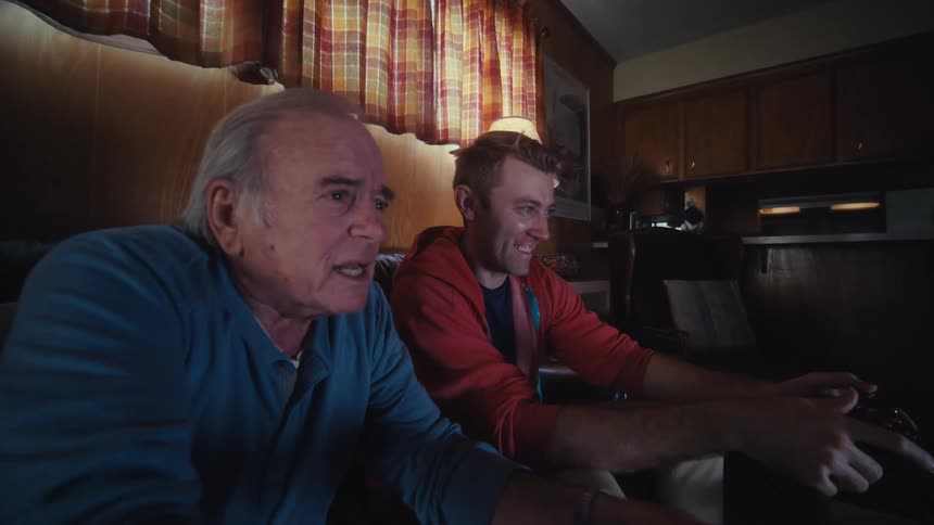 An old man and a young man are sitting on a couch in a living room and are playing video games using controllers