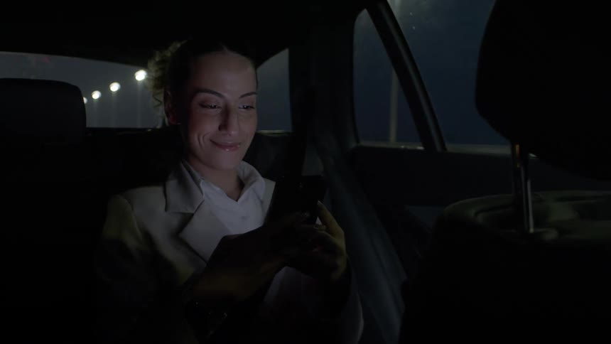 A womans face with a subtle smile as she is using her cell phone while sitting in the back seat of a car