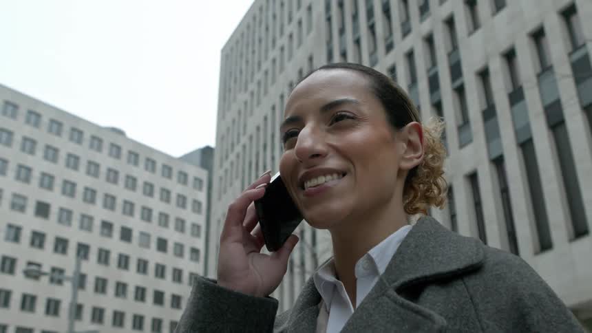 A woman with curly hair is talking on a smartphone while standing in front of a tall multistory office building