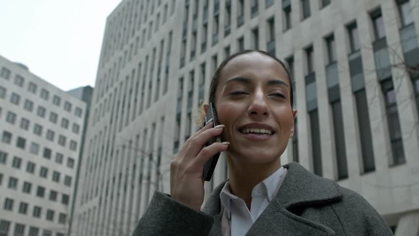 A woman talking on the phone and is smiling while looking ahead and she is wearing a gray coat and behind her there are some buildings