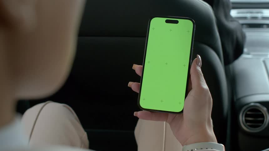 A woman sits in the back of a car holding a smartphone with a green screen in her hand