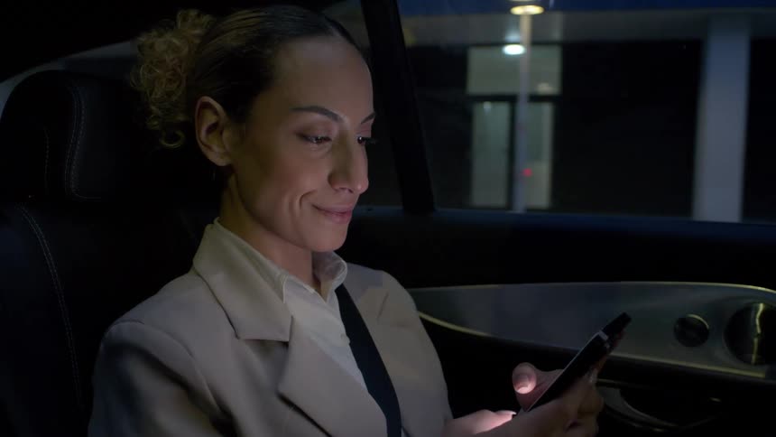 Woman riding through the city in a rear passenger seat at night smiles while scrolling on her smartphone