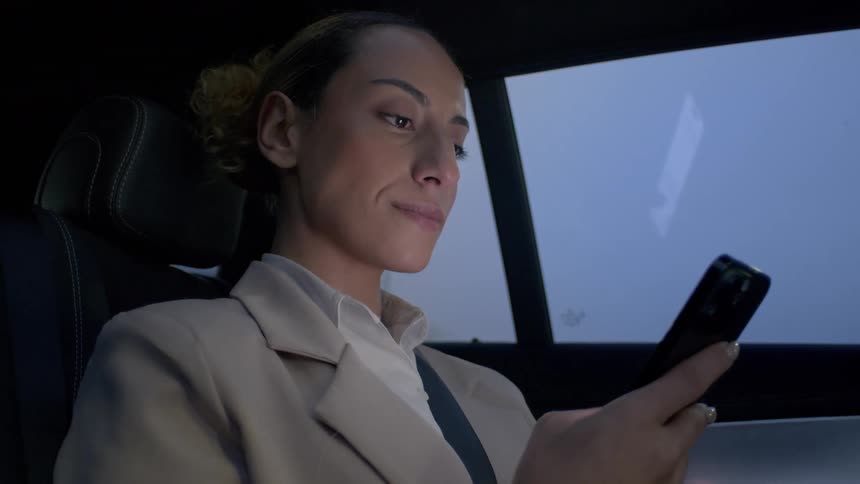 A woman with a subtle smile is looking at her cell phone while sitting in the back seat of a car and then looks out the window