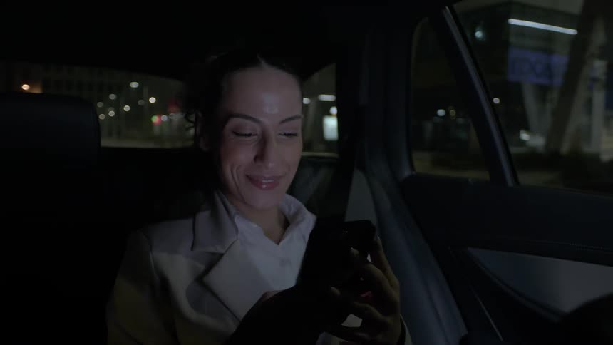 A woman sits in the backseat of a car at night looking at her phone