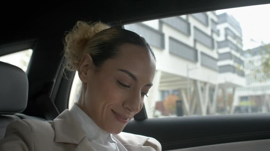 A woman with curly hair tied in a bun sits inside a car with the door open preparing to exit