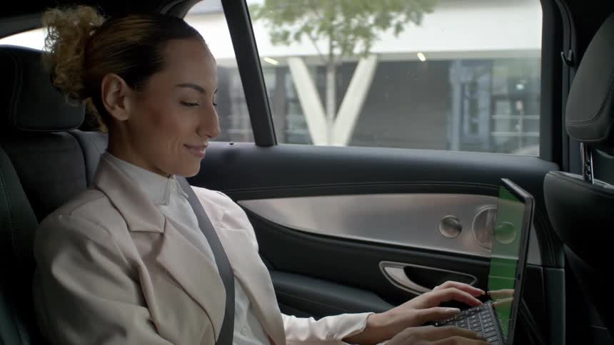 A woman is sitting in the backseat of a car working on a laptop