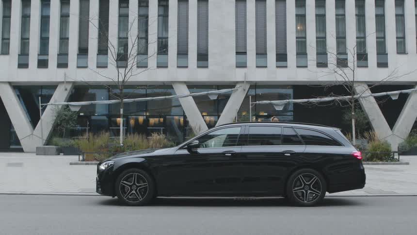 A woman stands on a sidewalk in front of a modern building waiting for a black taxi