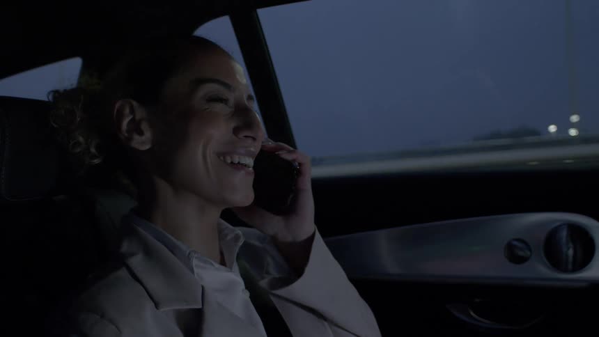 A woman is sitting in the backseat of a car at night talking on a smartphone with the car window showing a dark view of the outside