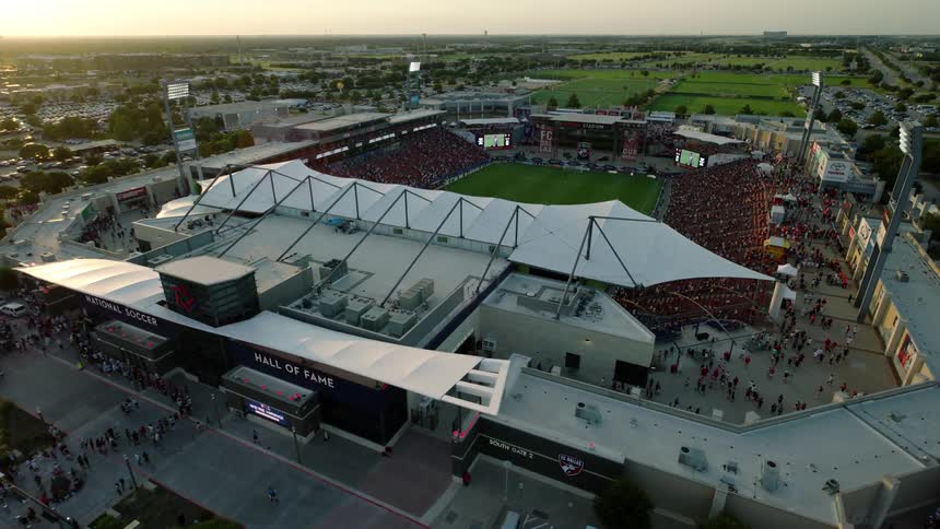 A soccer stadium packed with people with surrounding areas including a parking lot and other buildings