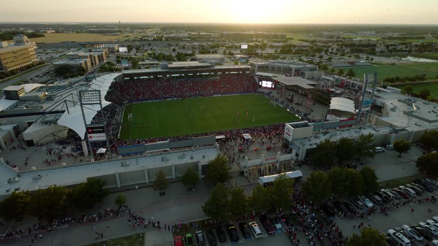 A soccer stadium filled with spectators with a large field in the center and a surrounding parking lot with cars and people