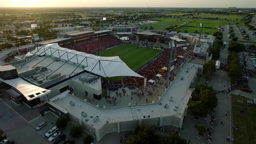An overhead shot shows a packed outdoor stadium with people entering and a game in progress