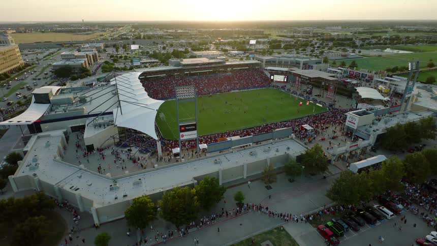 An aerial view of a stadium with a green lawn and stands packed with people with an outdoor parking lot full of cars