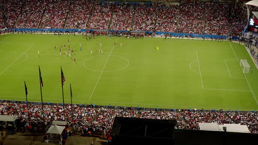 A large crowd watches a soccer match in a stadium with players positioned on the field