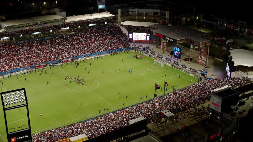 A packed soccer stadium at night with a large crowd in the stands players and officials on the field
