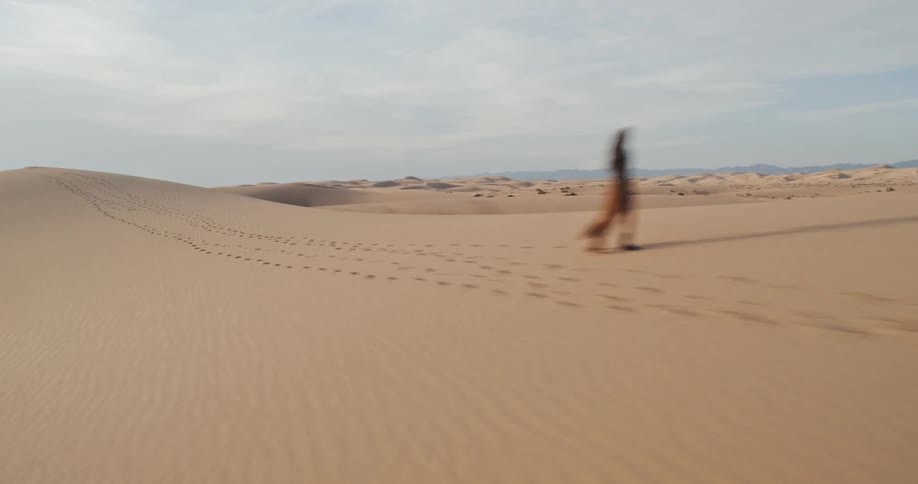 A person is walking alone in a vast desert leaving a trail of footprints in the sand under a partly cloudy sky