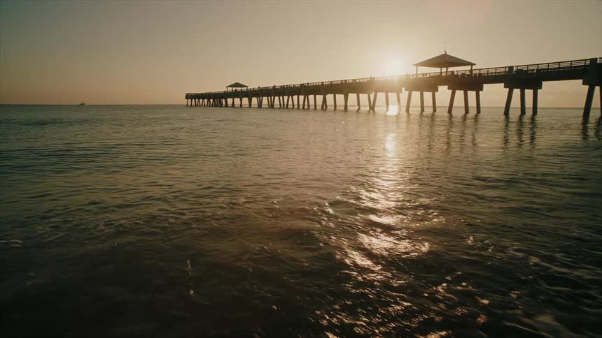 A long pier stretching into the calm ocean at sunset with the sun casting a warm glow on the water and the silhouettes of people enjoying the view