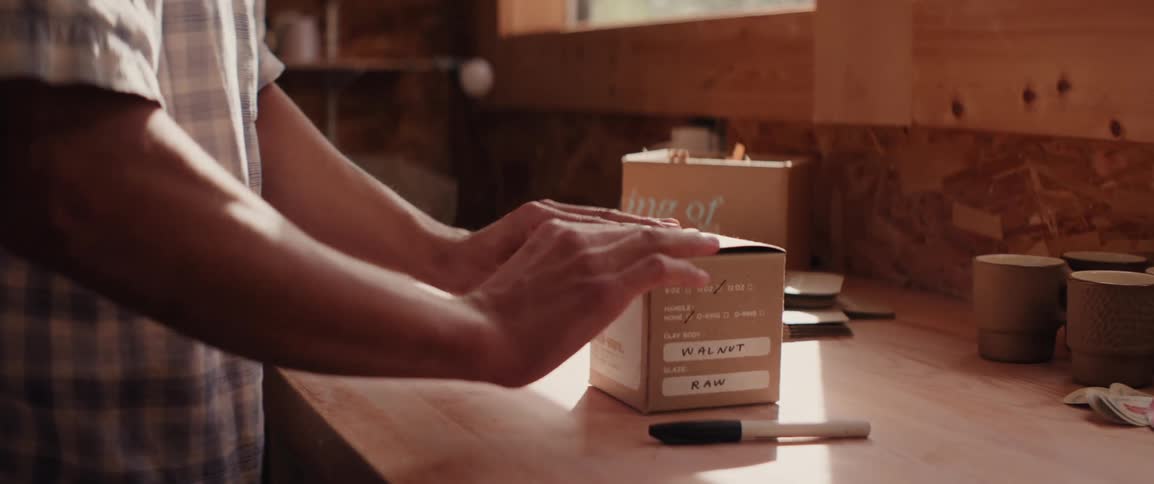 A man carefully closes a cardboard box filled with a finished ceramic mug in preparation of shipping