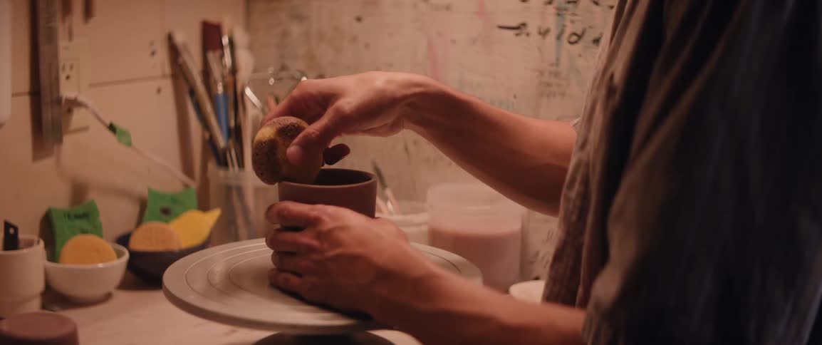 A potters hands carefully shape a piece of clay on a pottery wheel with other ceramic creations visible in the background