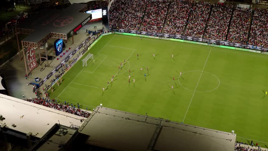 An aerial view captures a nighttime soccer game at toyota stadium showcasing a packed crowd a brightly illuminated field and dynamic player action with the ball moving swiftly across the pitch