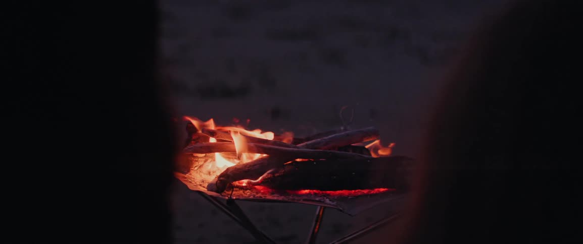 A small campfire crackles and glows with warm light on the beach at dusk