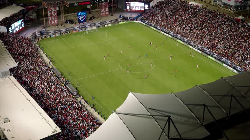 A night time shot of a soccer game at a stadium showing players on the field and a large crowd in the stands