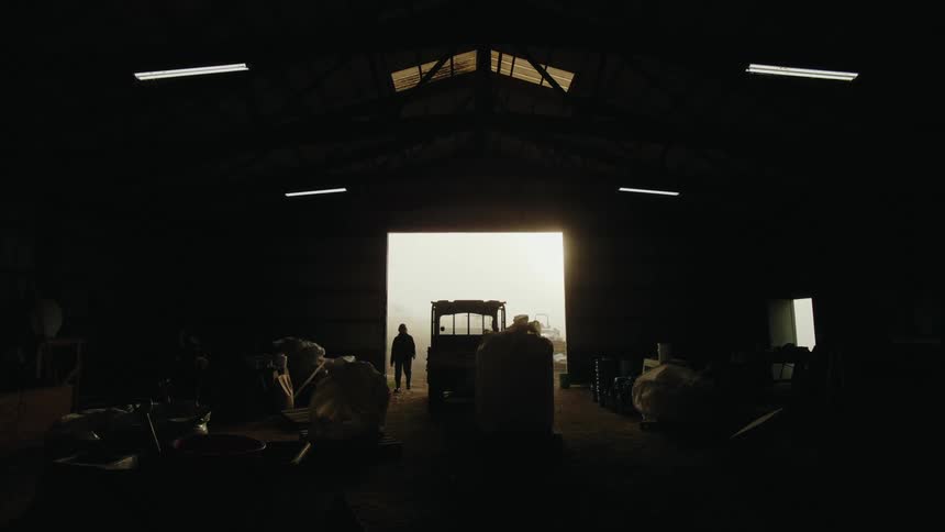 The silhouette of a person walking toward a parked tractor inside a dimly lit barn is visible against the bright light of the doorway