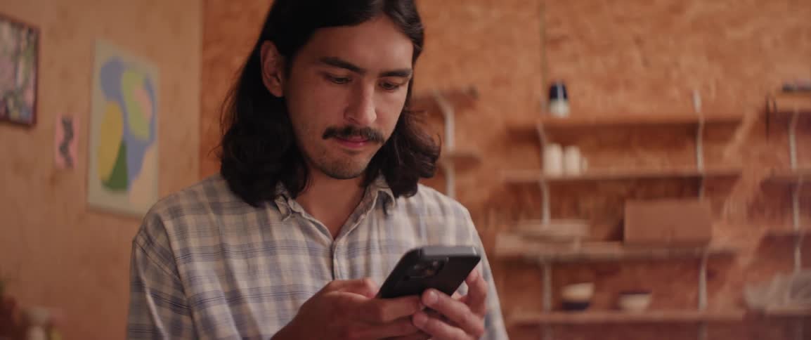 A person wearing a plaid shirt is looking at a smartphone in a room with wooden walls and shelves