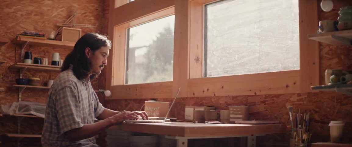 A man with long hair wearing a shirt sits at a desk working on a laptop in front of the windows