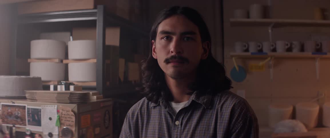 Portrait of a man with long hair and a mustache sitting in a pottery workshop looking directly at the camera