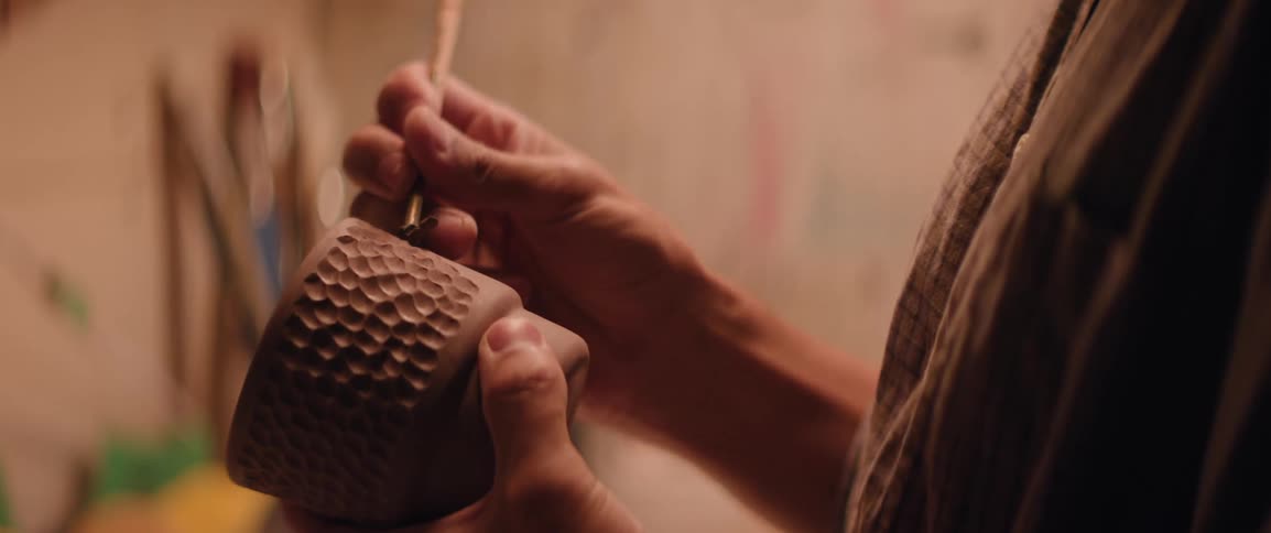A person is intricately carving a textured pattern into a clay pot using a small tool in a workshop setting