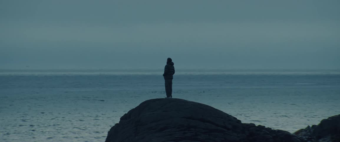 The silhouette of a person standing on a rock facing the sea under stormy skies