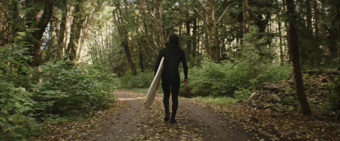 A person wearing a black wetsuit is walking down a forest path carrying a surfboard under one arm surrounded by dense green foliage and tall trees
