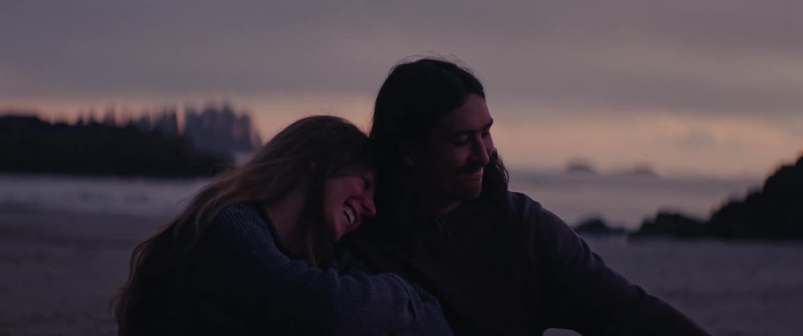 Two people sit closely together on the beach at dusk and one rests their head on the others shoulder