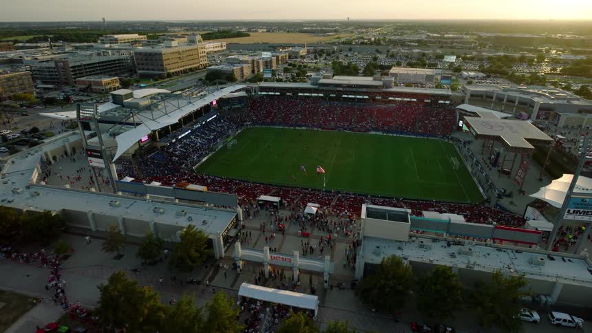 A soccer game happening at dusk with a packed stadium lit up and players running around all set against the backdrop of a city with buildings and lights visible in the distance