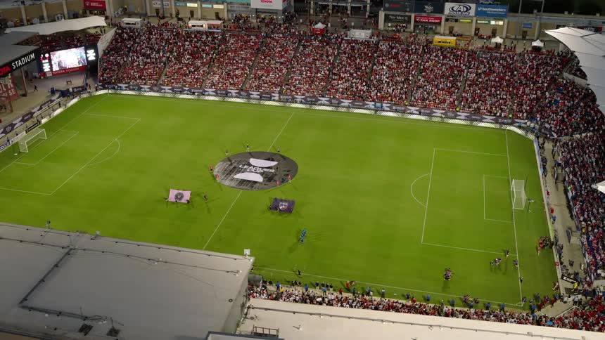 A packed soccer stadium at night with a large crowd in the stands players and officials on the field