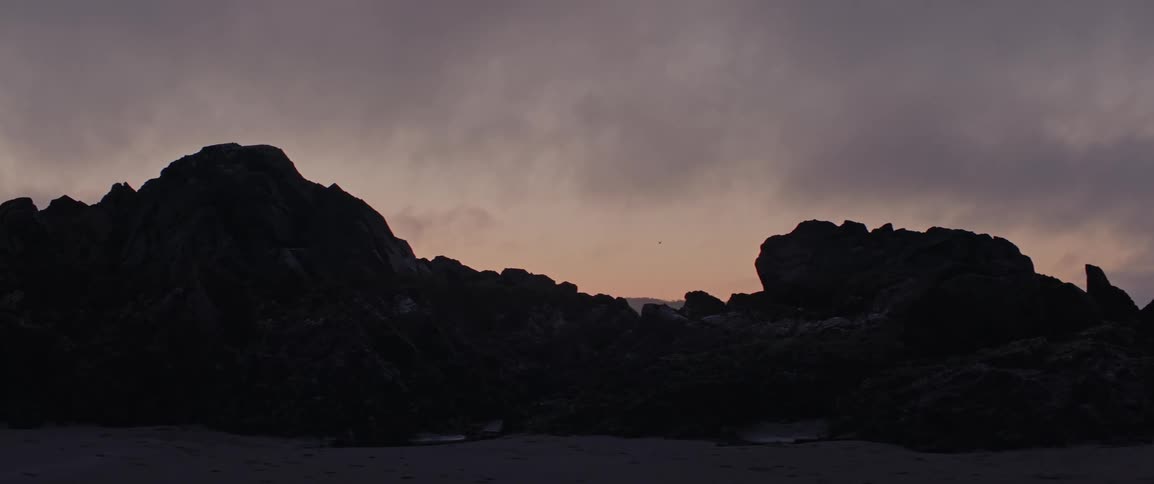 A huge dark rock mass on a beach after the sun has set and dusk sets in