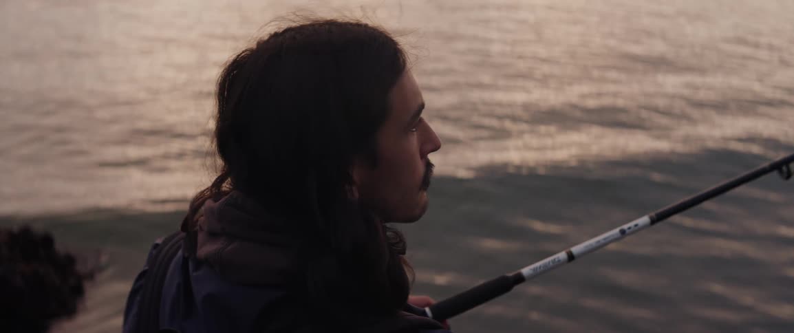 Close up of a man with long hair and a mustache fishing in the ocean at sunset