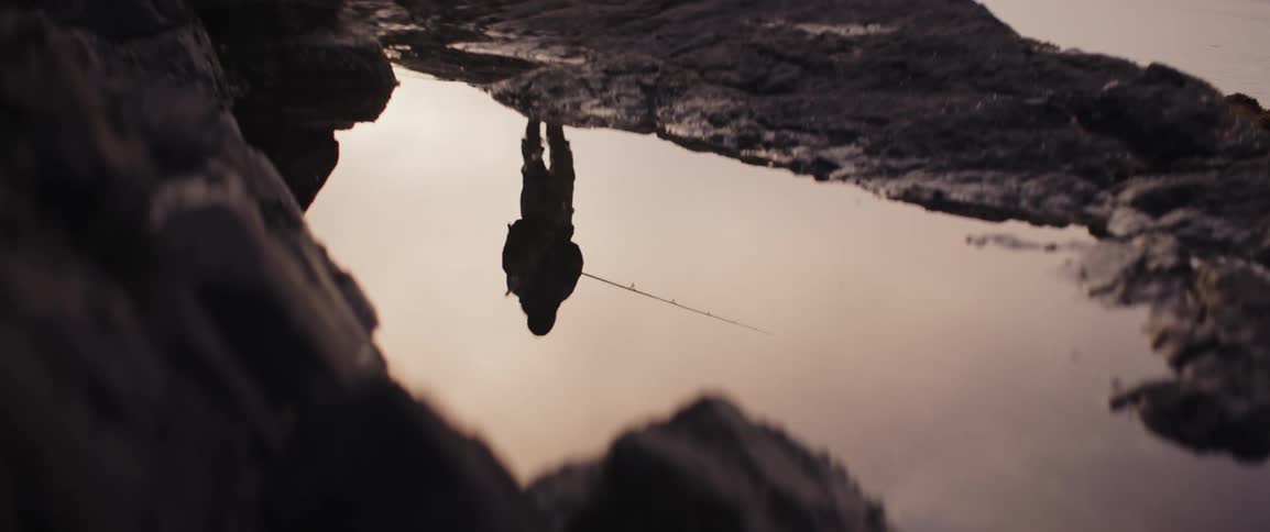 The water reflects a person standing on a rock fishing into the ocean at sunset